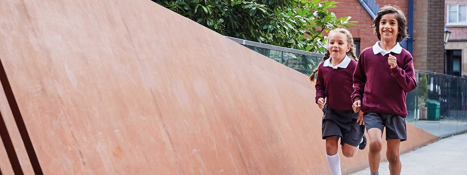 A primary school aged girl and boy wearing white polo shirts with red sweatshirts and grey shorts running happily outside