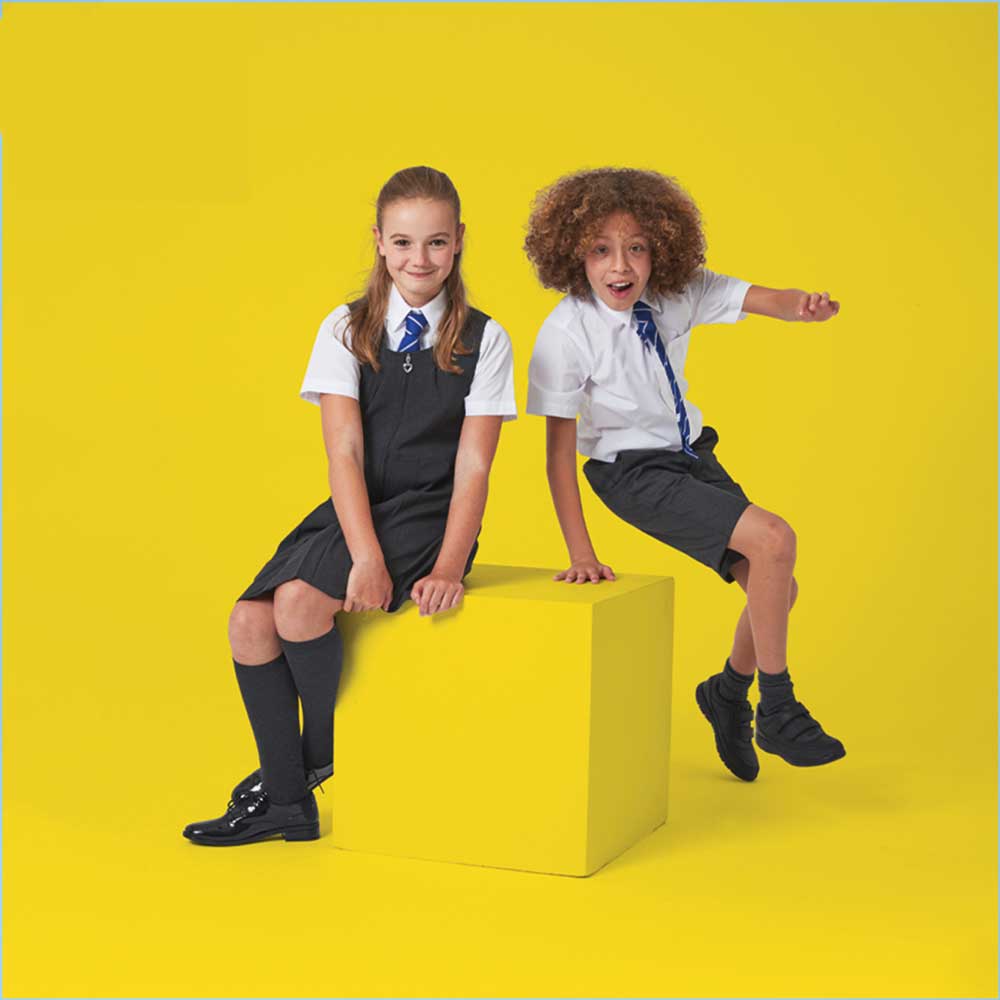 2 primary school children, a boy and a girl, in school uniform sat on a yellow box with a yellow background