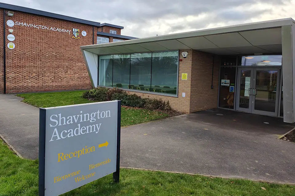 Shavington Academy exterior showing a brick building with a modern glass entrance canopy and a gray reception sign in the foreground on grass.