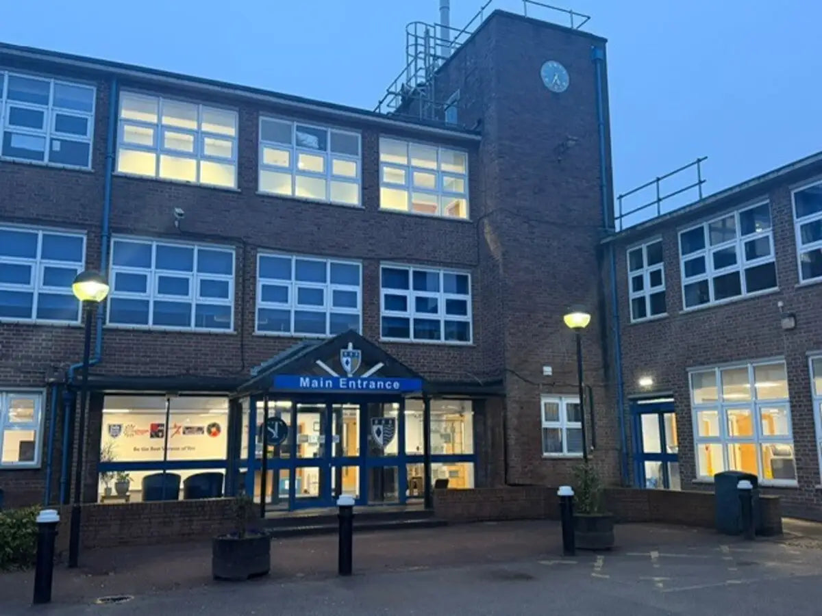 The Weaverham High School building with a main entrance canopy labeled 'Main Entrance,' multiple lit windows, blue-gray sky, and street lamps in the foreground.